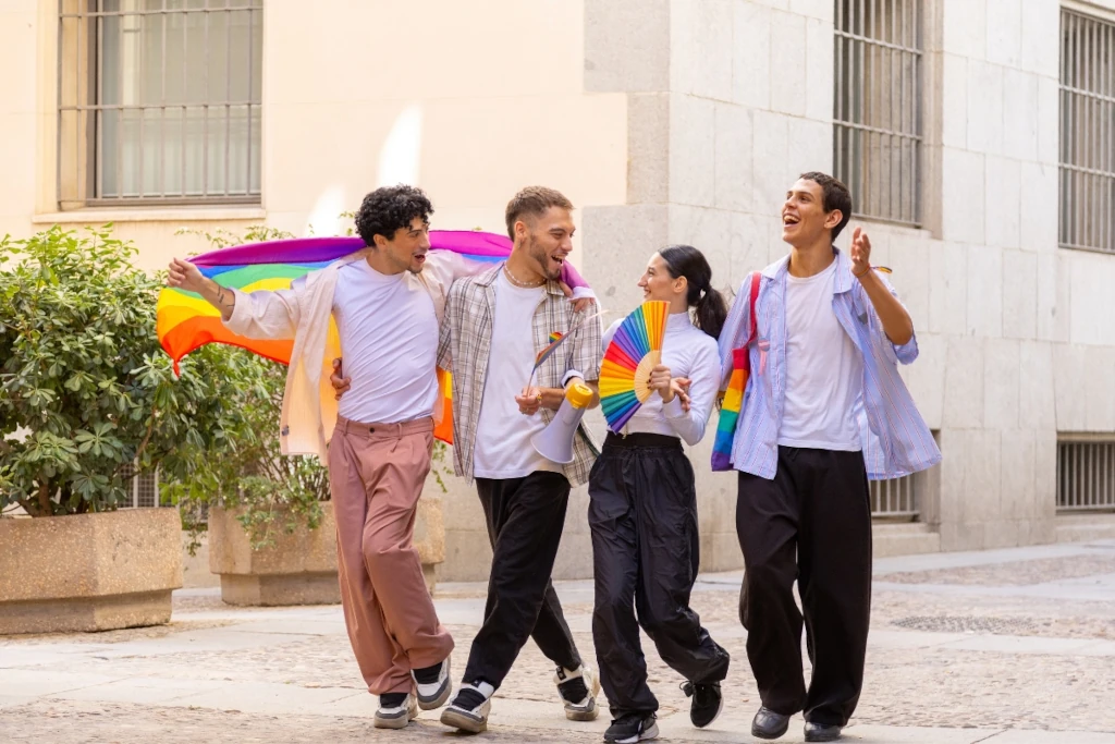Vier Freund*innen feiern gemeinsam Pride in der Stadt – mit Regenbogenflagge, Fächer und Megafon